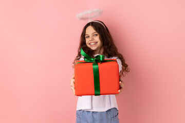 Portrait of joyful cheerful little girl wearing white T-shirt and nimb over head, holding out wrapped present box, congratulating with holiday. Indoor studio shot isolated on pink background.