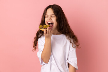 Sugar addiction. Portrait of satisfied little girl wearing white T-shirt biting delicious donut, looking with desire to eat sweet dessert. Indoor studio shot isolated on pink background.