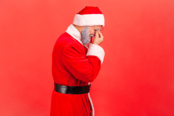 Unhappy elderly man with gray beard wearing santa claus costume crying, expressing sadness, feeling desperate sorrow, suffering depression. Indoor studio shot isolated on red background.