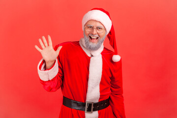 Fototapeta na wymiar Hello! Happy elderly man with gray beard wearing santa claus costume raising palm to wave hi, greeting with hospitable friendly smile. Indoor studio shot isolated on red background.
