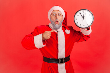 Fototapeta na wymiar Portrait of shocked elderly man with gray beard wearing santa claus costume pointing at wall clock waiting for appointment, keeping mouth open. Indoor studio shot isolated on red background.