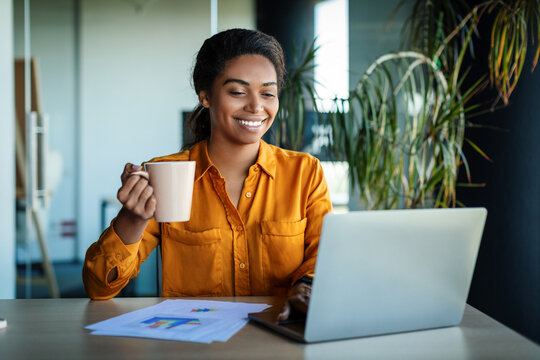 Happy african american businesswoman working on laptop and drinking coffee, sitting at workplace in office