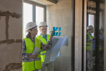 two engineers in protective vests and white helmets glue stickers on the window and discuss the construction plan of the facility or the scheme. European male and female engineers perform fire safety