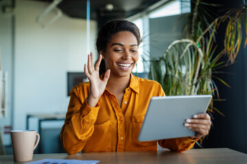 Happy black businesswoman sitting at table in office, having video chat with business partners,...