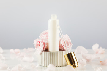 Obraz na płótnie Canvas White dropper bottle with serum on a marble podium on a white table with tender pink roses and rose petals. Natural beauty product based on rose flowers. Soft focus style
