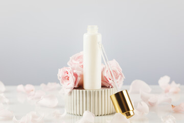 White dropper bottle with serum on a marble podium on a white table with tender pink roses and rose petals. Natural beauty product based on rose flowers. Soft focus style