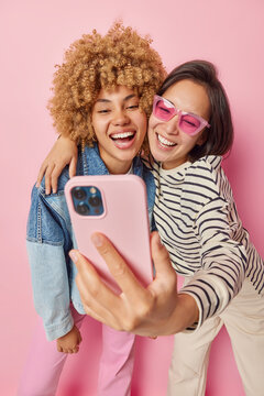 Vertical shot of happy diverse female friends embrace and have fun take selfie via smartphone smiles toothily dressed in casual clothes isolated pink background. Friendship and free time concept