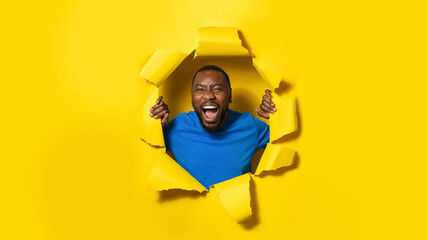 African american man shouting, reacting on something amazing posing through hole in yellow paper background, panorama