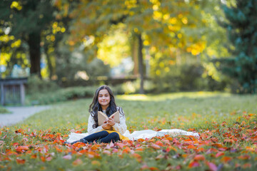 A pretty dark-haired 10-year-old girl in a knitted sweater reads a book sitting in a clearing in an autumn park. Picnic in nature. Happy child is learning.