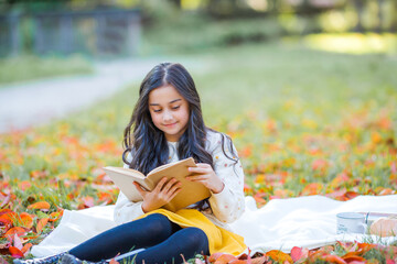 A pretty dark-haired 10-year-old girl in a knitted sweater reads a book sitting in a clearing in an...