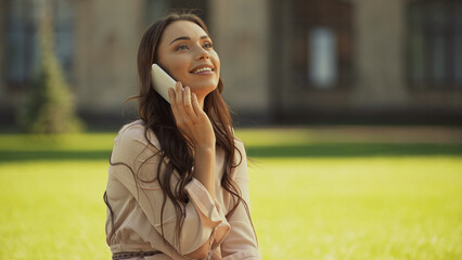Smiling brunette woman talking on smartphone in blurred park.