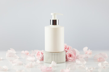 Mock up beauty product. White cosmetic dispenser bottle on a marble podium on a white table with tender pink roses and rose petals. Natural beauty product based on rose flowers. Soft focus style