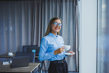 Image of a happy young woman in a classic shirt and glasses smiling and drinking coffee while standing by the window in a modern office with large windows. Remote work