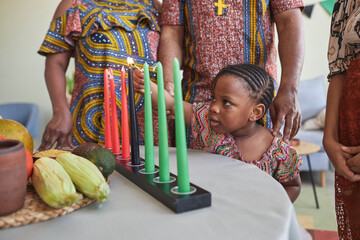 African little girl lighting candles on table while celebrating Kwanzaa holiday together with her...