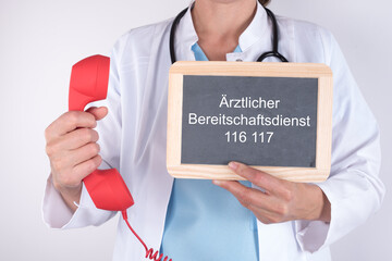Ärztin mit einem roten Notfalltelefon und einer Tafel auf der Ärztlicher Bereitschaftsdienst 116...