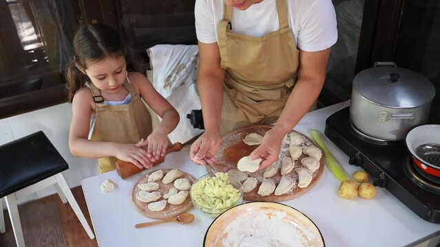 Top view of woman putting mashed potatoes on a rolled out dough and enjoying cooking dumplings with her cute daughter. Pretty little girl learns making vareniki, using rolling pin rolls out the dough