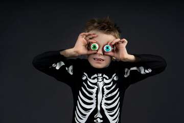 Cute little halloween boy with toy eyes looking at camera .wearing Halloween costume skeleton on dark background with copy space for text . Space for advertising