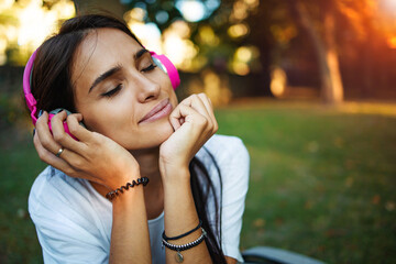 Beautiful young woman with headphones enjoy music outdoors. Girl repose or lie down at sunset on green grass.