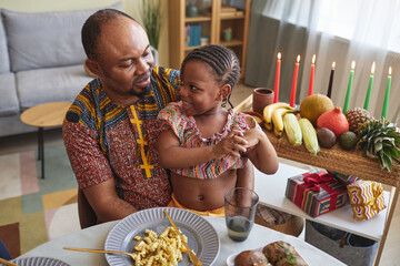 African dad talking to his little girl while she sitting on his knees during holiday dinner at table at home