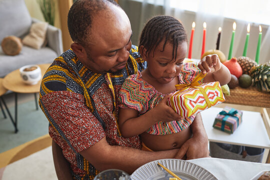 African little girl opening gift box while sitting on knees of her father at dining table