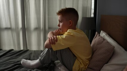 Side view of lonely sad boy sitting on bed with hugging knee and looking away, feeling pressure and problem. Unhappy stressed child resting alone in bedroom with negative emotion, slow motion.