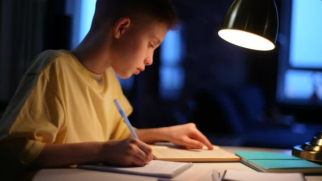 Close-up side view of redhead school boy pupil studying at home sitting at table under light of lamp. Smart cute Caucasian school student writing in exercise book doing homework, learning at desk.