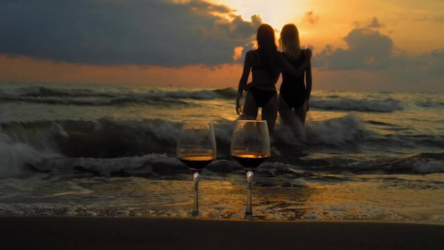 Romantic evening with stunning sunset, two women entering scream at sunset, against backdrop of two glasses of red wine and sea waves. Beautiful landscape on vacation or resort. Pair.