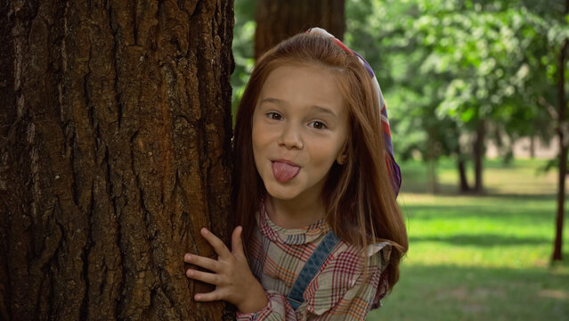 Red haired girl sticking out tongue near tree in park.