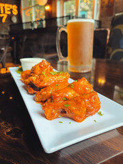 Plated hot steamy saucy buffalo chicken wings with ranch and a chilled frosted mug full of beer with foam on a dark wood bar restaurant table with window lighting - vertical