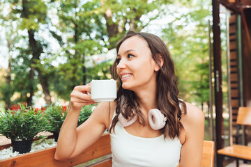 A young beautiful woman with headphones is sitting at a table in a summer cafe and drinking coffee or tea. generation z.