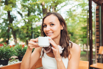 A young beautiful woman with headphones is sitting at a table in a summer cafe and drinking coffee or tea. generation z.