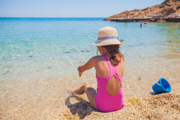Child with hat have fun at sand beach by sea water. Back view. Beautiful seascape view, turquoise color water sea scenic. Sunny day. Summer vacation.