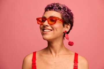 Fashion portrait of a woman with a short haircut in colored sunglasses with unusual accessories with earrings smiling on a pink bright background