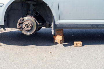 Fragment of a silver car without a wheel. The support is a column of old bricks. There is an asphalt road surface. Background. Close-up.