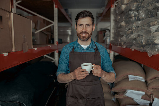 Smiling barista in uniform drinking coffee on warehouse with bags of coffee beans 