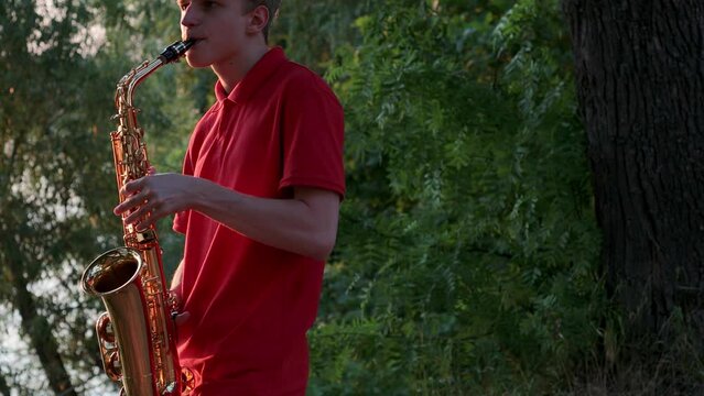 A young guy plays the saxophone near the river bank at sunset. Close-up.