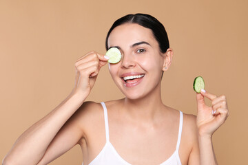 Obraz na płótnie Canvas Beautiful young woman putting slices of cucumber on eyes against beige background