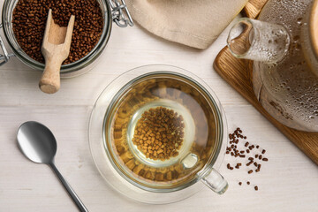Cup with buckwheat tea, hot teapot and granules on white wooden table, flat lay