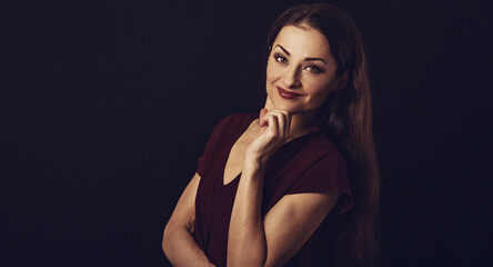 Fototapeta na wymiar Beautiful young business toothy smiling woman thinking and looking happy in burgundy blouse and with red lipstick on dark shadow black background with empty copy space. Portrait