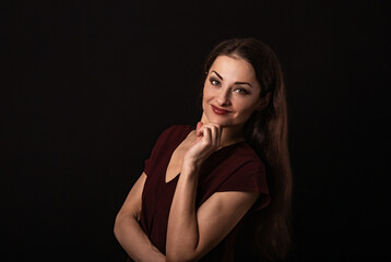 Fototapeta premium Beautiful young business toothy smiling woman thinking and looking happy in burgundy blouse and with red lipstick on dark shadow black background with empty copy space. Portrait
