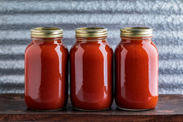Three jars of homemade water bath canned crushed tomato sauce made from homegrown tomatoes on a wooden table and industrial background - 528584590