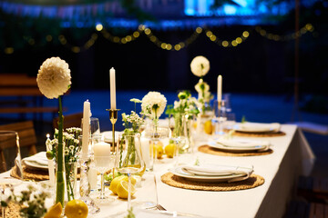 Part of long festive table decorated with candles and white dahlia flowers in restaurant lit by dark blue light creating romantic atmosphere