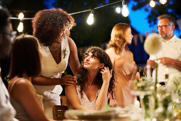 Two happy young brides in white attire looking at one another during conversation among guests gathered for wedding party
