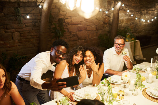 Two lesbian brides boasting with their wedding rings by served festive table while looking at smartphone screen held by young black man