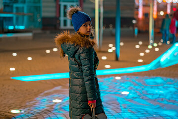 A Caucasian young girl in blue winter clothes stands on an illuminated street. Town. Tree. Nightfall. Eve. Activity. Cute. Fashion. Season. Cinematic. Illuminated. Lighted. Luminous. Colors. Wind