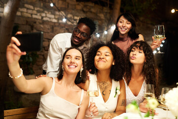 Young cheerful friends communicating in video chat or taking selfie by festive table while enjoying wedding party in restaurant