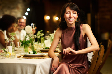 Young elegant brunette woman in silk evening gown sitting by served table and looking at camera while enjoying wedding party
