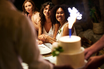 Happy young lesbian couple looking at big white wedding cake with sparkler while sitting by festive table among their guests