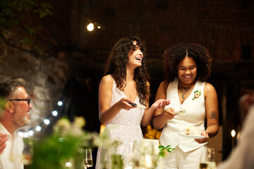 Cheerful young lesbian brides in white wedding attire spreading pieces of cut big cake among guests...