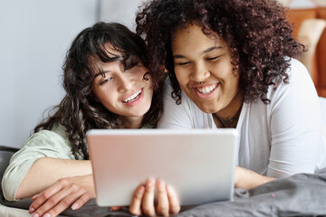 Two cheerful girlfriends with tablet communicating in video chat or choosing online goods while relaxing in bed in the morning